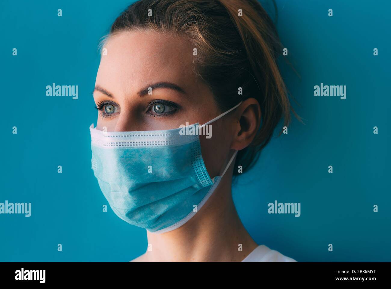 Portrait of young female doctor in medical mask and white gown on blue ...